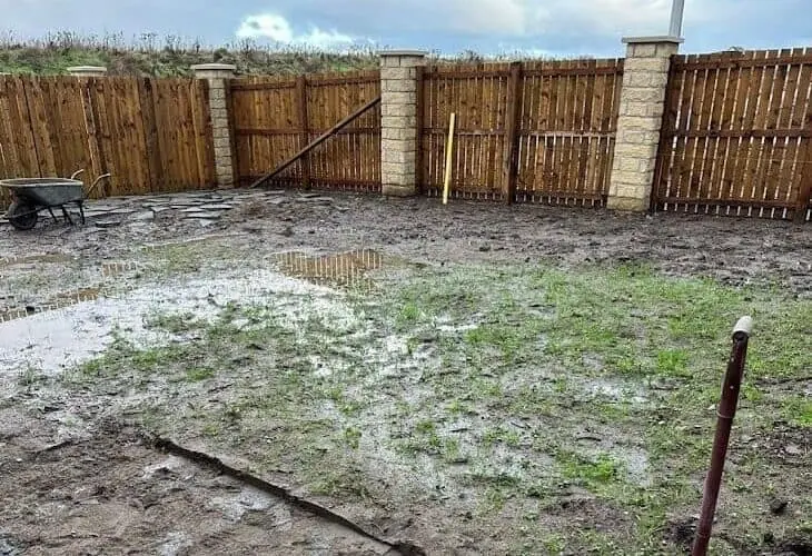 Muddy, waterlogged back garden with puddles in front of a timber fence and stone pillars