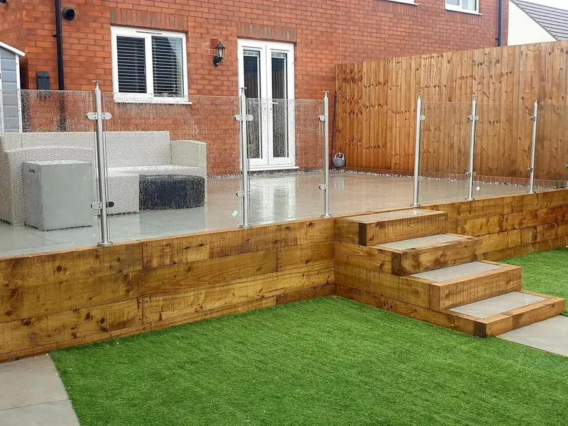 Raised deck with glass balustrade and timber sleeper steps leading down to an artificial lawn