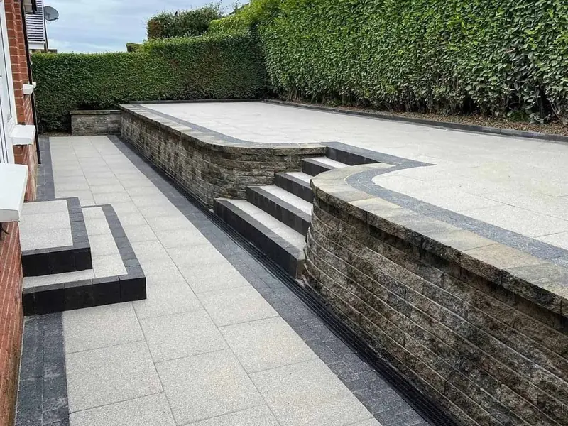 Stone-effect garden wall with built-in steps and a pale paved terrace above a hedge-lined garden