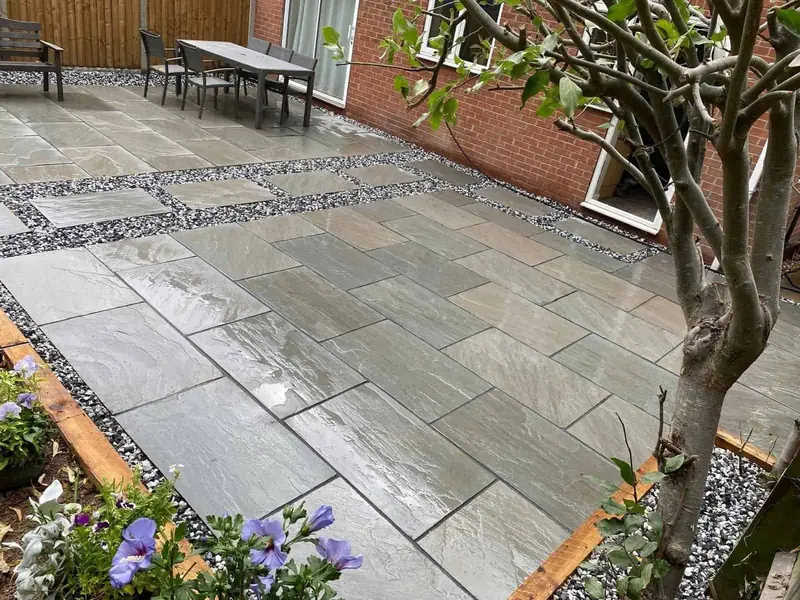 Grey paved patio with bands of dark gravel, timber-edged planting borders and an outdoor dining table